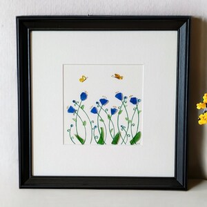 May include: A framed artwork featuring a blue flower arrangement with green stems and leaves. Two yellow butterflies are flying above the flowers. The artwork is set against a white background.