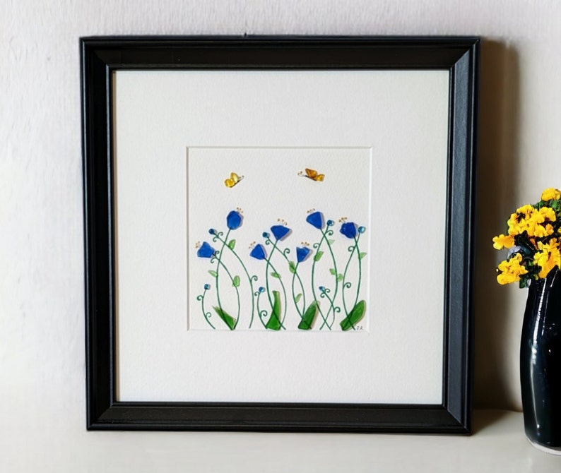 May include: A framed artwork featuring a blue flower arrangement with green stems and leaves. Two yellow butterflies are flying above the flowers. The artwork is set against a white background.