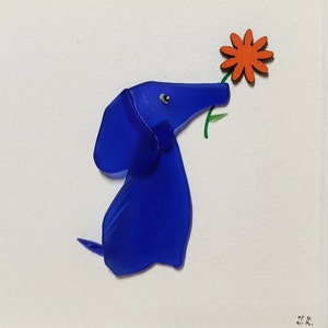 May include: A blue glass dachshund figurine holding a red flower. The figurine is on a white background with the artist's signature in the bottom right corner.