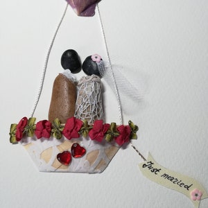 May include: A wedding themed craft made with seashells, stones, and fabric. A bride and groom are in a hot air balloon made of a seashell with a red and green garland around the basket. The balloon has a banner that says "Just married".