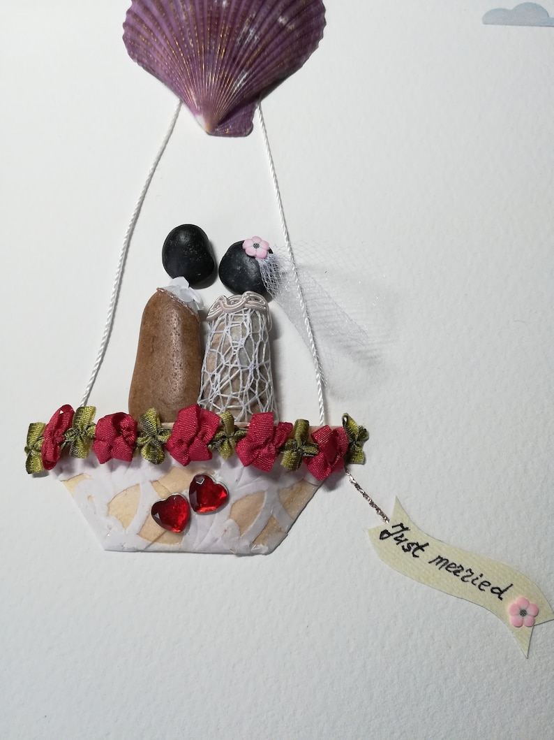 May include: A wedding themed craft made with seashells, stones, and fabric. A bride and groom are in a hot air balloon made of a seashell with a red and green garland around the basket. The balloon has a banner that says "Just married".