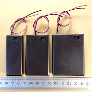 May include: Three black battery holders with red and black wires attached. The holders are labelled "ON-OFF". A ruler is below the holders, showing measurements in centimetres.