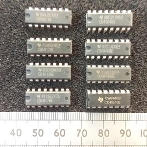 Op de afbeelding: Een verzameling van tien geïntegreerde schakelchips, elk met een rechthoekig grijs lichaam en meerdere pinnen. De chips zijn gelabeld met verschillende codes en nummers, waaronder "CD4001BE" en "CD4051BE". Een liniaal met centimeter markeringen.