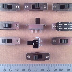 May include: An assortment of silver-colored toggle switches with black levers, arranged on a light brown surface. The switches vary in design, with some having mounting holes. A ruler with millimeter markings is at the bottom.
