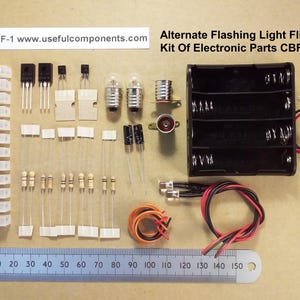 Op de afbeelding: Een verzameling elektronische componenten, waaronder transistors, weerstanden en een batterijhouder, gerangschikt op een lichtbruin oppervlak. De kit bevat een liniaal met cm-metingen en de tekst "Alternate Flashing Light Flip-Flop Kit Of Electronic Parts CBFF-1".