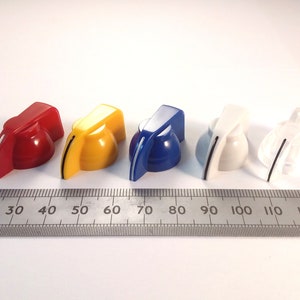 以下が含まれることがあります： 赤、黄、青、白、透明の5つのプラスチックノブ。ノブは先のとがった上部と平らな底部を持っています。白い表面に並べて配置され、その下に定規があります。