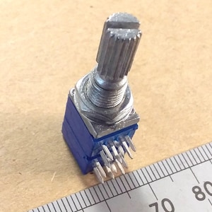 May include: A silver-colored potentiometer with a ridged shaft and a blue base. The electronic component has multiple pins and is positioned near a ruler, suggesting a measurement context.