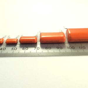 May include: An assortment of orange rectangular capacitors of varying sizes, arranged in a row. Each capacitor has two silver leads. A ruler with inch markings is placed beneath the capacitors, providing a size reference.