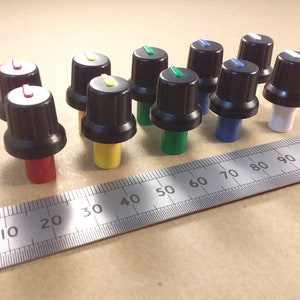 Peut inclure: Assortiment de 10 boutons de potentiomètre noirs avec des anneaux de couleur. Les boutons sont disposés en rangée sur une règle graduée en centimètres. Les couleurs des anneaux sont rouge, jaune, vert, bleu et blanc.