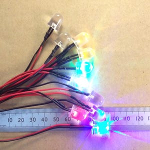May include: A selection of multi-coloured LED lights with clear plastic domes, connected by red and black wires. A ruler with centimetre markings is placed next to the lights. The lights are emitting various colours, including red, green, and blue.