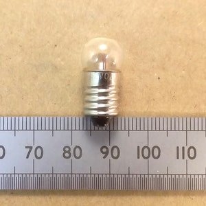 May include: A miniature clear glass light bulb with a silver base. The bulb is next to a ruler, providing a size reference. The filament is visible inside the glass. The base has the text "VO".