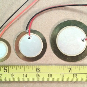 May include: Three round, gold-coloured, piezo-electric sound sensors with wires attached. The sensors are 3.8 cm, 5.1 cm, and 6.4 cm in diameter. The sensors are on a brown surface with a yellow measuring tape.