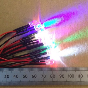 May include: A set of 10 multi-coloured LED lights with black and red wires. The lights are arranged in a row and are illuminated, casting a rainbow of colours on the surface below. A ruler is visible at the bottom of the image, with markings from 10 to 130.