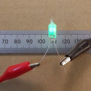 May include: A small, glowing green light bulb is connected to red and black crocodile clips. A ruler with centimetre markings is in the background. The light bulb is lit, indicating an electrical circuit.