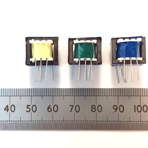 May include: Three small electronic transformers with different colored tape: yellow, green, and blue. Each transformer has a dark brown frame and several silver pins extending from the bottom. A ruler is placed below the transformers, with measurements in inches.