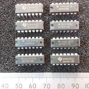 May include: A collection of integrated circuit chips, each with a rectangular body and multiple pins. The chips are marked with alphanumeric codes and the Texas Instruments logo. A ruler with centimetre markings is in the foreground.