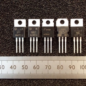 May include: Five electronic components with black tops and silver pins are arranged in a row. Each component has text, including model numbers like "317T" and "7805CT." A ruler with markings in inches is placed below the components.