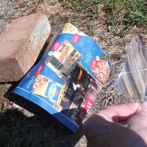 May include: A red-brown brick, a blue and white advertisement, and a feather are on the ground. The advertisement features images of food. The feather is held by a person.