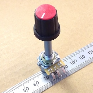 May include: A red knob with a white indicator line sits atop a black base, connected to a silver shaft and electronic component. A ruler with inch markings is in the background. The knob is a component for electronics projects.