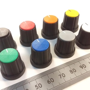 May include: A collection of eight black control knobs with colored tops, including red, orange, yellow, green, blue, gray, and white. A ruler with centimeter markings is in the foreground, indicating the size of the knobs.