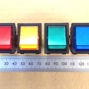 May include: Four illuminated rocker switches in red, yellow, green, and blue, each set in a black rectangular frame. A metal ruler with inch markings is placed below the switches, providing a size reference. The switches are likely for electrical or electronic applications.