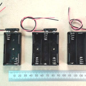 Op de afbeelding: Drie zwarte batterijhouders met rode en zwarte draden. De houders zijn ontworpen voor één, twee en vier batterijen. Een liniaal eronder toont metingen in centimeters, tot 15 cm.