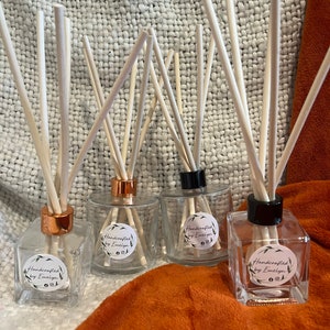 May include: Three glass bottles with wooden diffuser sticks and labels that say "Handcrafted by Emelyn". The bottles have copper and black caps. The bottles are sitting on a white and orange textured surface.
