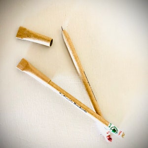 May include: Two beige pens with the text "Co-Simple Products" and a pen cap are displayed on a white surface. One pen is uncapped, revealing the writing tip. The pens are made of a light brown material, possibly recycled paper.