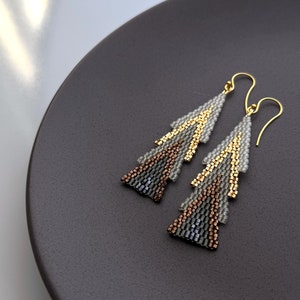 Può includere: Un paio di orecchini dorati con un design geometrico. Gli orecchini sono realizzati con piccole perline in tonalità di grigio, marrone e oro.