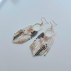 Op de afbeelding: Een paar gouden creolen met witte, zwarte en roze kralen franjes. De oorbellen zijn gemaakt van kleine, ronde kralen en hebben een geometrisch patroon.