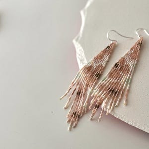 May include: A pair of long, beaded earrings with a pink, white and brown colour scheme. The earrings are made of small, round beads that are strung together in a fringe-like design.