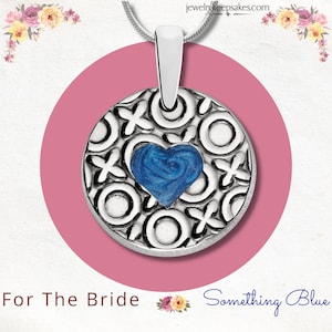 Puede incluir: Collar con colgante de plata con un dije de corazón azul. El corazón está rodeado por un círculo de plata con un patrón decorativo. El texto "Something Blue" es visible en la imagen.