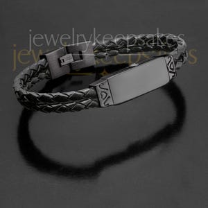 May include: A black braided leather bracelet featuring a rectangular metal ID plate with decorative etched scrollwork on the sides. The bracelet includes a matching black metal clasp mechanism and a smooth, polished center surface for engraving.