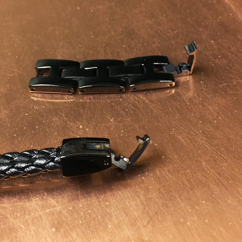 May include: Close-up of a black leather watch band with a black metal clasp. The clasp is open and the band is lying on a copper-colored surface.