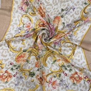 May include: A square scarf with a cream-colored background featuring a leopard print pattern. The scarf is adorned with floral and gold baroque designs. The flowers are in shades of orange, pink, and purple, with a tan border.