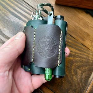 May include: A dark green leather multi-tool organizer featuring a dark brown leather front panel with a circular embossed landscape design of a sun over mountains. The organizer has light green stitching and holds a bright green metal tool.