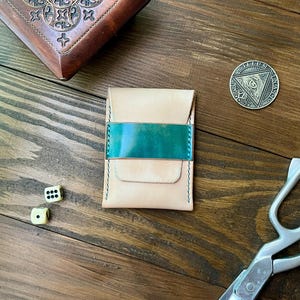 Puede incluir: Un tarjetero rectangular de cuero con acabado en beige natural y una banda horizontal en color verde azulado que contrasta en el centro. El artículo presenta costuras visibles en verde azulado a los lados y un diseño de solapa plegada.