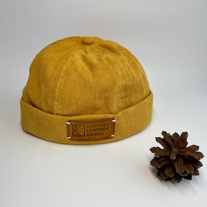 May include: Mustard yellow corduroy beanie hat with a rolled brim. A small leather tag with the text "KARTCEV LEATHER GOODS" is sewn on the brim. A pine cone sits to the right of the hat.