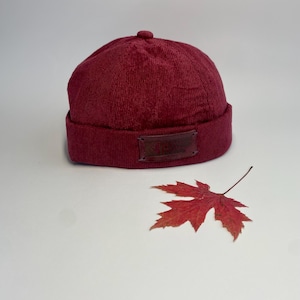 May include: A burgundy beanie hat with a small, round top detail and a brown leather patch. A red maple leaf rests beside the hat on a white surface. The hat has a ribbed brim and a textured fabric.