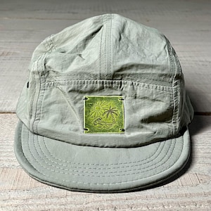 Pode incluir: Boné verde sálvia de cinco painéis com aba curva. O boné apresenta um patch quadrado verde com uma ilustração de sol, palmeira e água. Feito de um material leve, tem um design estruturado.
