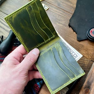 Op de afbeelding: Een bi-fold portemonnee gemaakt van olijfgroen leer met een verweerde afwerking, voorzien van meerdere kaartsleuven aan de binnenzijde en een biljettenvak met een biljet van vijftig dollar. De randen zijn gestikt met lichtgroene draad.