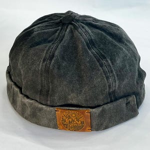 May include: A charcoal grey brimless cap featuring a six-panel construction with a button at the crown. A rectangular tan leather patch is stitched to the front cuff, displaying a laser-engraved illustration of a sun, mountains, and trees.
