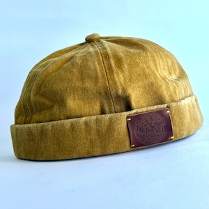Könnte beinhalten: Senffarbene, randlose Mütze mit kleinem, rundem Oberteil. Ein braunes Lederpatch mit Berg- und Sonnendesign ist vorne angebracht. Die Mütze ist aus einem strukturierten Stoff gefertigt.