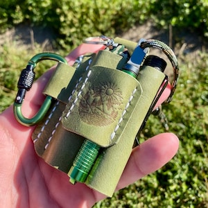 Puede incluir: Funda de transporte de cuero verde oliva con mosquetón verde. La funda contiene una pequeña linterna verde y un cuchillo plateado y negro. La funda tiene un diseño en relieve de sol y paisaje y costuras blancas.