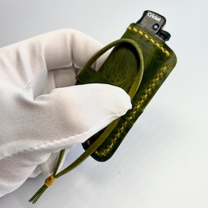 May include: A green leather lighter case with yellow stitching and a loop handle. The lighter is black with the word "Cricket" printed on it. The case is held by a gloved hand.