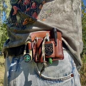 May include: A brown leather belt pouch with a compass, a small flashlight, and a fire starter. The pouch has a metal badge that reads "Adventure Awaits". The person is wearing a gray t-shirt with a mushroom graphic and blue jeans.
