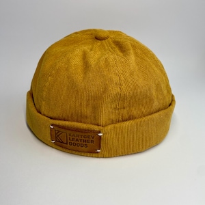 May include: Mustard yellow corduroy beanie hat with a folded brim. A small, rectangular leather patch with the text "KARTCEV LEATHER GOODS" is sewn onto the brim. The hat is made of a ribbed fabric and has a rounded crown.