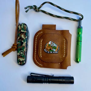Op de afbeelding: Een verzameling outdoor accessoires: een camouflage Zwitsers zakmes, een bruin leren etui met een "Back to the Woods" pin, een groene metalen tool en een zwarte zaklamp. De items zijn op een witte ondergrond geplaatst.