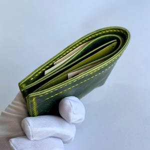 以下が含まれることがあります： 緑色の革製財布で、ステッチが見え、複数のコンパートメントがあります。財布は開いた状態で、内部が見えます。革は使い古したような外観で、ステッチは明るい緑色です。