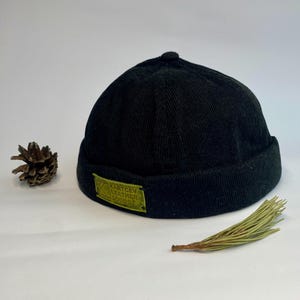 Peut inclure: Bonnet en velours côtelé noir avec un petit bord. Un patch rectangulaire vert avec le texte "KARTCEV LEATHER GOODS" est cousu sur le devant. Une pomme de pin et une brindille de verdure sont à côté du chapeau.